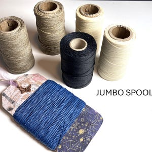 May include: Five cylindrical spools of thread in beige, tan, and black, alongside a flat rectangular card wrapped in blue thread. The card features a vintage-style print with a butterfly charm and the text JUMBO SPOOLS printed on the side.