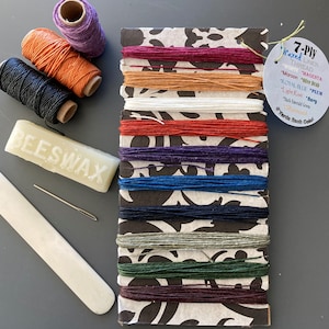 May include: A set of 7-ply waxed linen thread spools in various colors, including white, magenta, maroon, olive drab, royal blue, plum, light rust, navy, and dark emerald green. The spools are arranged vertically on a black and white patterned card. The card also includes a label with the text "7-Ply Waxed Linen Thread" and a list of the thread colors. There is also a bar of beeswax, a needle, and a wooden tool.