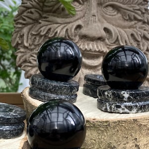 May include: Three polished black spheres, each resting on a dark speckled stone base. The spheres reflect the surrounding environment. The background features a stone carving of a face with foliage.