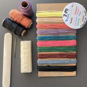 May include: A craft kit with spools of colored thread, a needle, beeswax, and a bone folder. The thread colors include black, salmon, lavender, green, and denim. A tag indicates the thread is 7-ply waxed linen.