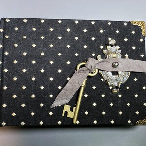May include: A black journal with a diamond pattern and brass corner protectors. A decorative brass key and clasp are attached with a gray ribbon. The journal's cover is black with a repeating pattern of small, gold diamonds.