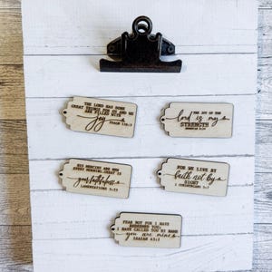 May include: A white wooden clipboard with a black metal clip featuring five rectangular light wood tags. Each tag is engraved with different religious verses including Psalm 126:3, Nehemiah 8:10, Lamentations 3:23, 2 Corinthians 5:7, and Isaiah 43:1.