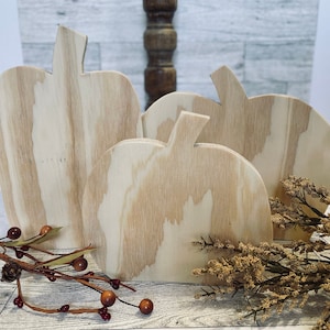 May include: Four unfinished wooden pumpkin cutouts in various sizes. The pumpkins have a natural wood grain and are arranged in a group. Decorative fall foliage with red berries and dried flowers are in the foreground. Perfect for fall decor.
