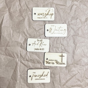 May include: Five rectangular wooden tags with laser-etched text and a small hole. The tags feature phrases like "worship," "In the Waiting," "Be still And Know," "He is Risen," and "It is finished," along with corresponding Bible verses.