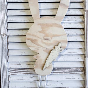 May include: Wooden bunny-shaped wall decor with a carrot. The bunny is made of light-colored wood and has long ears and a round face. A wooden carrot is attached to the bunny's body. The bunny has two dangling strings with wooden beads at the end.