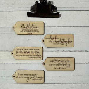 May include: Wooden tags with inspirational quotes and Bible verses. The tags are light brown with dark brown text. The quotes include "God is love", "I have loved you with an everlasting love", and "We love because He first loved us". A metal clip is at the top.