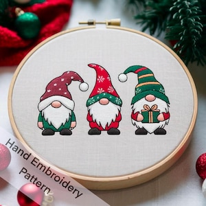 以下が含まれることがあります： 木製のフレームに入った、3体のクリスマスのノームの手刺繍パターン。各ノームは白いひげ、赤または緑の衣装、雪の結晶またはストライプのデザインの尖った帽子をしています。1体のノームはプレゼントを持っています。