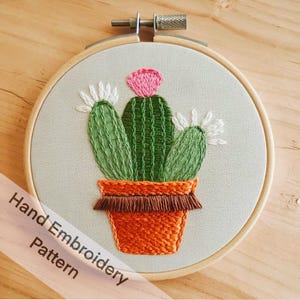 Op de afbeelding: Handborduurpatroon van een cactusplant in een terracotta pot met een bruine franje. De cactus is groen met een roze bloem en witte accenten.