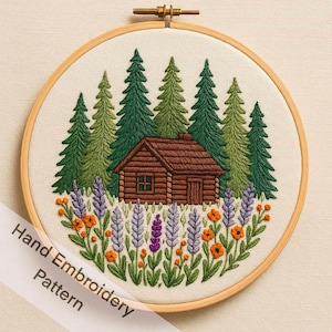 Op de afbeelding: Handgeborduurde hutscène in een houten hoepel. Het ontwerp toont een bruine hut genesteld tussen groene en geelgroene dennenbomen, met paarse en oranje bloemen op de voorgrond. De tekst "Hand Embroidery Pattern" staat op een wit lint.