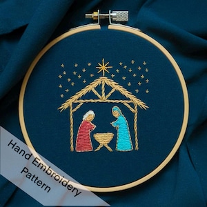 May include: Hand embroidery pattern of the nativity scene with a gold star, a wooden manger, and two figures in red and blue standing inside. The embroidery is on a dark blue fabric.