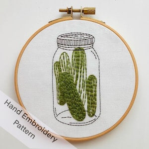 May include: Hand-stitched embroidery art featuring a jar of pickles. The design is on white fabric, framed by a wooden hoop. The pickles are green with detailed stitching, and the jar is outlined in black thread. The text "Hand Embroidery Pattern" is on a white label.