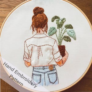 Op de afbeelding: Handborduurpatroon met een vrouw met bruin haar in een knot, die een wit shirt en een blauwe spijkerbroek draagt en een potplant met groene bladeren vasthoudt. Het patroon is op een witte stoffen achtergrond.