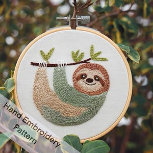 Op de afbeelding: Handborduurpatroon van een luiaard die aan een tak hangt. De luiaard is groen en bruin met een glimlachend gezicht. Het patroon is in een houten borduurring.