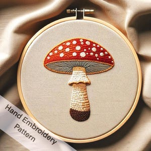 May include: A hand-embroidered pattern of a red and white mushroom with a gray cap and a brown stem. The mushroom is stitched on a beige fabric in a wooden embroidery hoop.