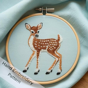 May include: Hand embroidery pattern featuring a brown and white spotted fawn with a light blue background. The pattern is in a wooden embroidery hoop.