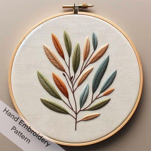 Może przedstawiać: Wzór haftu ręcznego przedstawiający gałąź z liśćmi w odcieniach zieleni, błękitu, pomarańczy i brązu. Haft jest prezentowany w drewnianym obręczy do haftu.