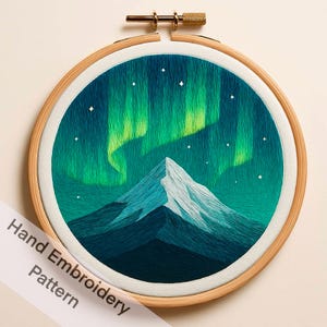 May include: Hand-embroidered hoop art featuring a mountain scene under the aurora borealis. The design showcases a mountain peak in shades of blue and white, with green and teal aurora lights and white stars. The embroidery is set in a wooden hoop.