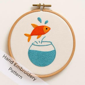 以下が含まれることがあります： 手刺繍のフープアート。オレンジ色の金魚が青いボウルから飛び跳ねています。デザインはクリーム色の生地に施され、木製のフープで縁取られています。「Hand Embroidery Pattern」の文字が斜めのバナーにあります。