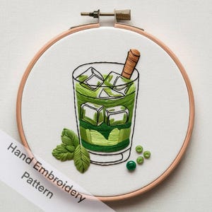 May include: Hand embroidery pattern of a green iced drink with a straw in a glass, surrounded by green leaves and green beads on a white background.