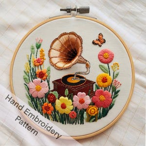 May include: Hand embroidery pattern featuring a vintage gramophone with a butterfly and flowers in shades of pink, yellow, orange, and green.