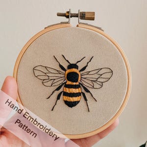 May include: A hand embroidery pattern featuring a bee with black and yellow stripes and translucent wings. The bee is stitched on a beige fabric and framed in a wooden hoop.