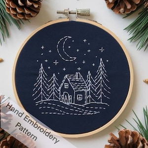 May include: Hand-stitched embroidery art on navy blue fabric, featuring a white crescent moon, stars, a house, and pine trees. The design is set in a wooden embroidery hoop. The text "Hand Embroidery Pattern" is visible.