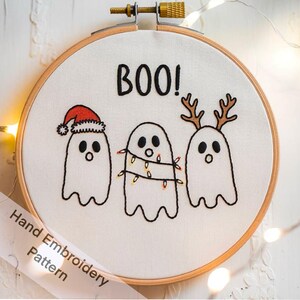 May include: Hand-stitched embroidery hoop art featuring three cartoon ghosts. One ghost wears a Santa hat, another is wrapped in Christmas lights, and the third has antlers. The word "BOO!" is stitched above the ghosts. The hoop is made of wood.