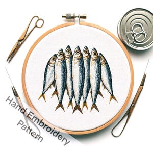 May include: Hand-stitched embroidery pattern of sardines in a wooden hoop. The sardines are detailed with blue, silver, and yellow thread. A metal can, scissors, and needles are placed around the hoop. The text "Hand Embroidery Pattern" is visible.