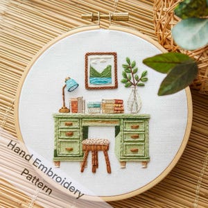 Op de afbeelding: Een handgestikt borduurpatroon van een groene bureau met laden, een kruk, een lamp, boeken, een vaas met groen en een ingelijst beeld van een landschap. De borduurwerk is tentoongesteld in een houten ring.