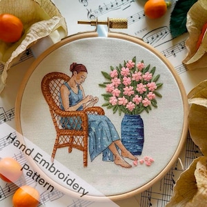 May include: Hand embroidery pattern featuring a woman sitting in a wicker chair with a bouquet of pink flowers in a blue vase. The woman is wearing a blue dress and has brown hair. The embroidery is done on a white fabric and is framed in a wooden hoop.