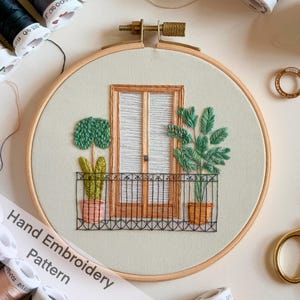 Op de afbeelding: Een met de hand geborduurd hoepelkunstwerk dat een balkon met een deur, twee potplanten en een smeedijzeren leuning toont. De borduurwerk is uitgevoerd in tinten groen, bruin en zwart op een lichtgroene stof.