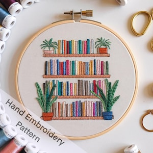 May include: A hand-stitched embroidery hoop with a design of a bookshelf filled with colorful books and potted plants. The text "Hand Embroidery Pattern" is written below the hoop.