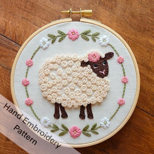 May include: A white sheep with brown eyes and a pink flower on its head is embroidered on a white fabric in a wooden embroidery hoop. The sheep is surrounded by a wreath of green leaves and pink and white flowers.