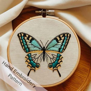以下が含まれることがあります： 木製の刺繍枠に張られた、クリーム色の生地に手刺繍された蝶の模様。 蝶は、ターコイズ、黒、金色のディテールが特徴です。 「Hand Embroidery Pattern」の文字がリボンに表示されています。
