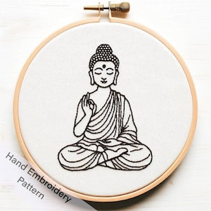 May include: Black and white line drawing of a meditating Buddha embroidered on white fabric in a wooden embroidery hoop.