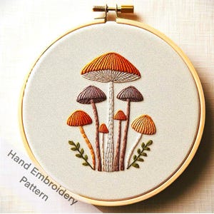 May include: Hand embroidery pattern featuring a group of mushrooms with orange, brown, and white colors. The mushrooms are embroidered on a white fabric and are surrounded by green leaves.