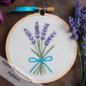 May include: A hand embroidery pattern featuring a bouquet of lavender flowers tied with a blue ribbon. The pattern is displayed in a wooden embroidery hoop.