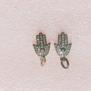 May include: Two silver-toned Hamsa hand charms. Each charm is encrusted with small, clear stones and features a small, round, pink stone in the center. The charms have a loop at the bottom for attachment.