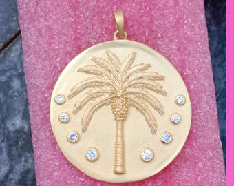 May include: Gold-toned pendant with a detailed palm tree design at the center. The pendant is circular, with small, clear gemstones encircling the tree. The pendant has a loop at the top for a chain.