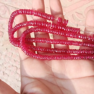 Può includere: Fili di perline di pietre preziose sfaccettate di colore rosso intenso. Le perline sono infilate insieme, formando più anelli. Le pietre preziose hanno un aspetto scintillante e il colore è un rosso ricco e vibrante. Le perline sono tenute in una mano.