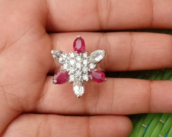 May include: A silver ring with a flower-like design. It features six oval red gemstones and six clear gemstones around a cluster of small, clear stones. The ring is set on a hand.