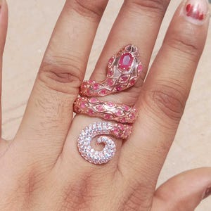 May include: A rose gold snake ring featuring a coiled design that wraps around the finger. The head is set with a central oval red stone, while the body is encrusted with numerous small pink stones and the tail features clear white stones.