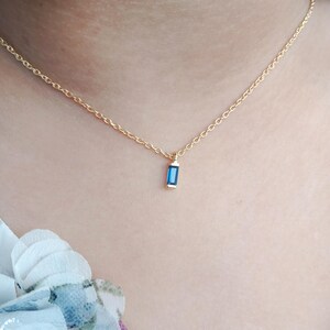 May include: A delicate gold necklace featuring a small, rectangular blue gemstone pendant. The pendant is set in a gold frame and hangs from a fine gold chain. The necklace is shown against a skin tone background.