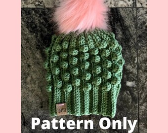 May include: A green crochet beanie with a pink faux fur pom pom. The beanie has a textured stitch pattern and a small label that says "TOASTY". The text "Pattern Only" is written below the beanie.