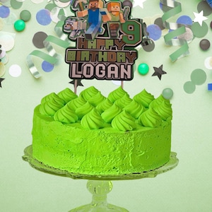 May include: A vibrant green birthday cake on a clear glass cake stand. The cake is decorated with green frosting swirls and topped with a Minecraft-themed cake topper that reads "Happy Birthday Logan" and the number 9.