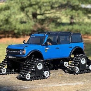 May include: A blue toy truck with black treads, designed for off-road use. The truck has a black roof and a detailed front grille. The wheels are replaced with black treads, and the truck is sitting on a wooden surface.