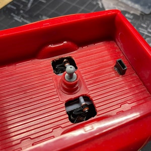 Op de afbeelding: Close-up van de rode laadbak van een speelgoed pick-up truck. De laadbak is gemaakt van plastic en heeft een getextureerd oppervlak. Er zijn twee gaten in de laadbak, elk met een zwarte metalen as en een zilveren metalen wiel.