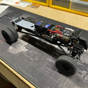Op de afbeelding: Een zwart en grijs RC-autochassis met een zwarte en grijze motor en een zwart en rode accu. De auto staat op een zwart en grijze kaart.