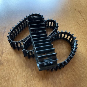Op de afbeelding: Drie zwarte plastic rupsbanden met een getextureerd oppervlak en een reeks tanden langs de randen.