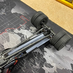 Op de afbeelding: Een close-up van een zwart speelgoedtruckchassis met twee zwarte banden. De truck staat op een zwart-grijze wereldkaart muismat.